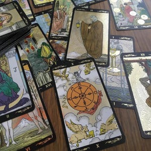 May include: A collection of tarot cards with gold accents and black borders. The cards feature detailed illustrations and symbols, including a wheel, figures, and celestial imagery. The cards are spread out on a wooden surface.