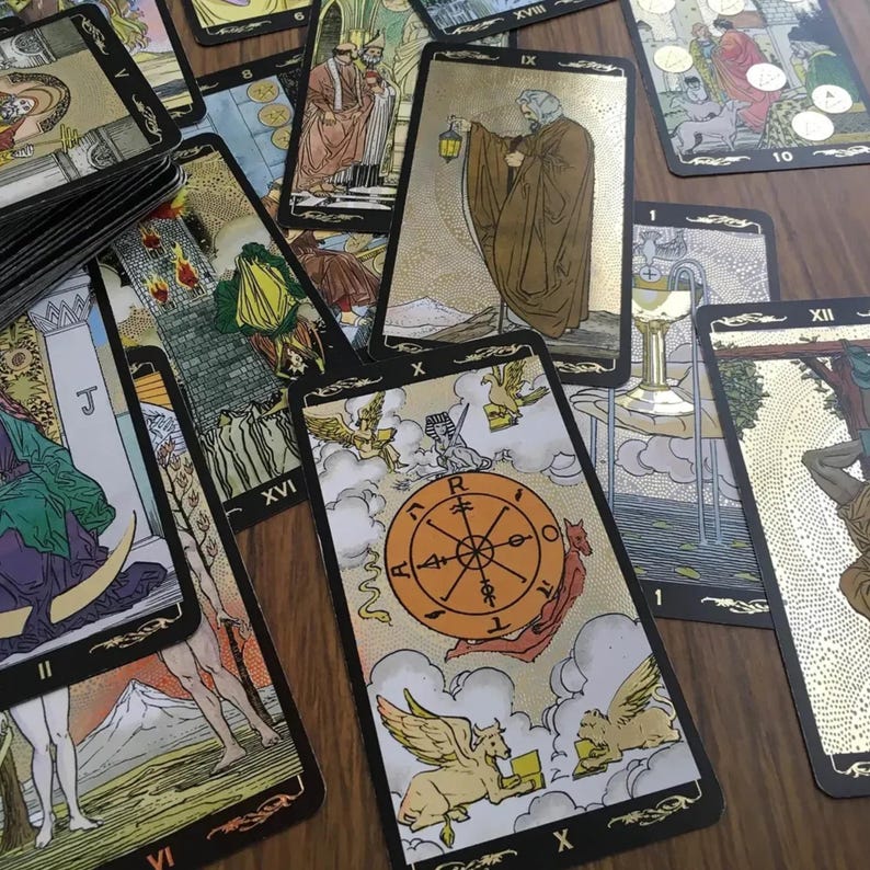 May include: A collection of tarot cards with gold accents and black borders. The cards feature detailed illustrations and symbols, including a wheel, figures, and celestial imagery. The cards are spread out on a wooden surface.