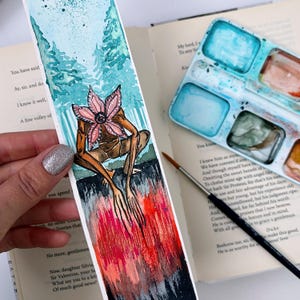 May include: Watercolor bookmark featuring a Demogorgon-inspired illustration. The artwork depicts the creature with a flower-like head, set against a backdrop of a forest and a fiery, abstract lower section. The bookmark is placed on an open book.