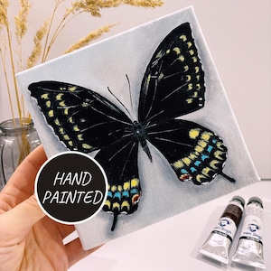 May include: Hand painted canvas featuring a black butterfly with yellow and blue accents on a white background. The canvas is labelled "Hand Painted".