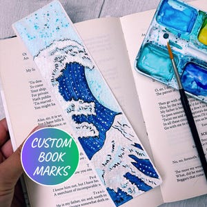 May include: Hand-painted bookmark featuring a blue and white wave design, with a watercolor palette and brush in the background. The bookmark has the text "CUSTOM BOOK MARKS" on it. The bookmark is placed on top of an open book.