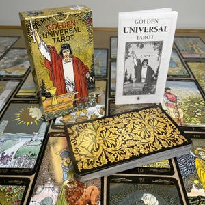 May include: A Golden Universal Tarot deck with a gold and black floral design on the card backs. The box and guidebook are visible, featuring illustrations of figures and the text "GOLDEN UNIVERSAL TAROT". The cards are spread out, showcasing various illustrations.