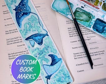 Peut inclure: Marque-page artisanal avec des illustrations de raies manta à l'aquarelle bleues et turquoise. Le marque-page porte l'inscription "CUSTOM BOOK MARKS" sur un autocollant. Une palette d'aquarelle et un pinceau sont également visibles.