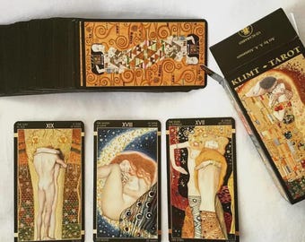 May include: A deck of Klimt Tarot cards featuring gold-toned, intricate artwork. The set includes a black storage box labeled KLIMT TAROT and three face-up cards displaying figures in artistic styles, including The Sun XIX, The Moon XVIII, and The Star XVII.