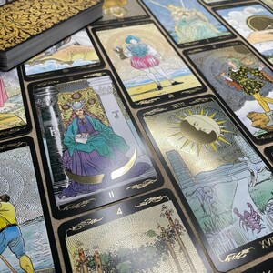 May include: A deck of tarot cards with gold foil accents and black borders. The cards feature detailed illustrations with figures, symbols, and numbers. A gold-embossed box is visible in the upper left corner. The cards are arranged on a neutral surface.