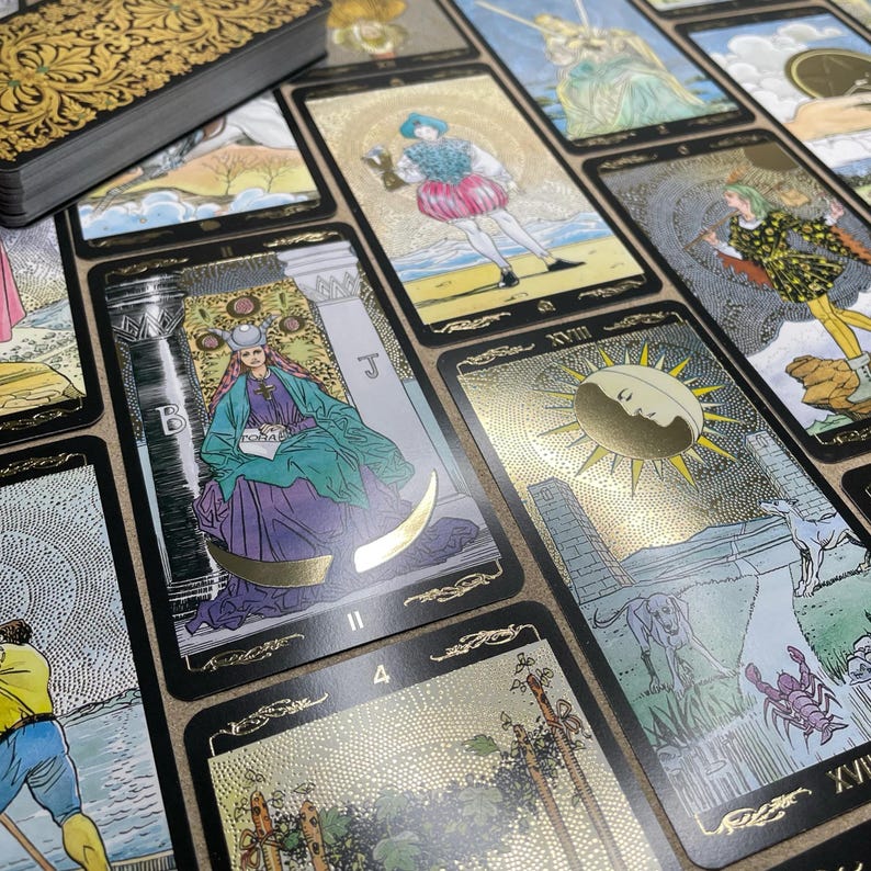 May include: A deck of tarot cards with gold foil accents and black borders. The cards feature detailed illustrations with figures, symbols, and numbers. A gold-embossed box is visible in the upper left corner. The cards are arranged on a neutral surface.