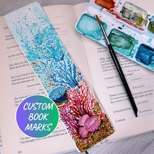May include: A hand-painted bookmark with a vibrant coral reef design in shades of blue, red, and pink. The bookmark is on an open book with the text "CUSTOM BOOK MARKS" on a circular sticker.