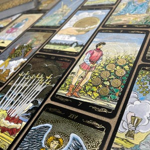May include: A collection of tarot cards with gold accents and black borders. The cards feature detailed illustrations, including figures, symbols, and numbers. The cards are arranged on a surface, showcasing various designs and themes.