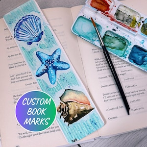Puede incluir: Un marcapáginas pintado a mano con un diseño de concha, estrella de mar y caracola. El marcapáginas es de color azul claro con detalles de acuarela y el texto "CUSTOM BOOK MARKS". Un pincel y una paleta de acuarela están en el fondo.