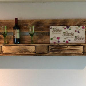 Könnte beinhalten: Ein rustikales Holzregal mit drei Fächern, mit einer Weinflasche, zwei Weingläsern und einem gerahmten Schild mit dem Text "Liebe jeden Moment, Lache jeden Tag, Liebe unendlich".