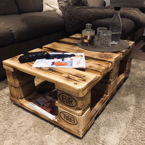 Puede incluir: Una mesa de centro de madera r&uacute;stica hecha de palets reutilizados. La mesa tiene un acabado de madera natural y presenta el texto "EUR EPAL" en el lateral. La mesa est&aacute; situada sobre una alfombra beige delante de un sof&aacute; marr&oacute;n.