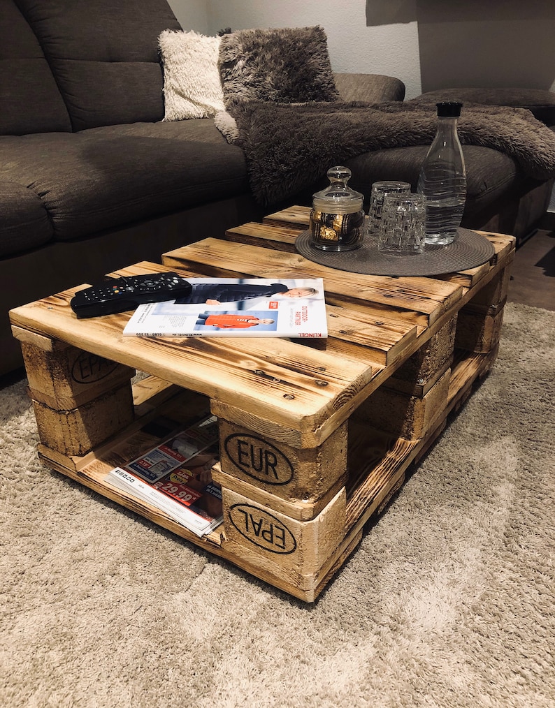 Puede incluir: Una mesa de centro de madera r&uacute;stica hecha de palets reutilizados. La mesa tiene un acabado de madera natural y presenta el texto "EUR EPAL" en el lateral. La mesa est&aacute; situada sobre una alfombra beige delante de un sof&aacute; marr&oacute;n.