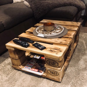 Puede incluir: Una mesa de centro de madera hecha de palets reutilizados. La mesa tiene un aspecto r&uacute;stico y desgastado y presenta las marcas "EUR" y "EPAL". Dos mandos a distancia est&aacute;n sobre la superficie superior de la mesa, y una revista est&aacute; metida en el estante inferior.