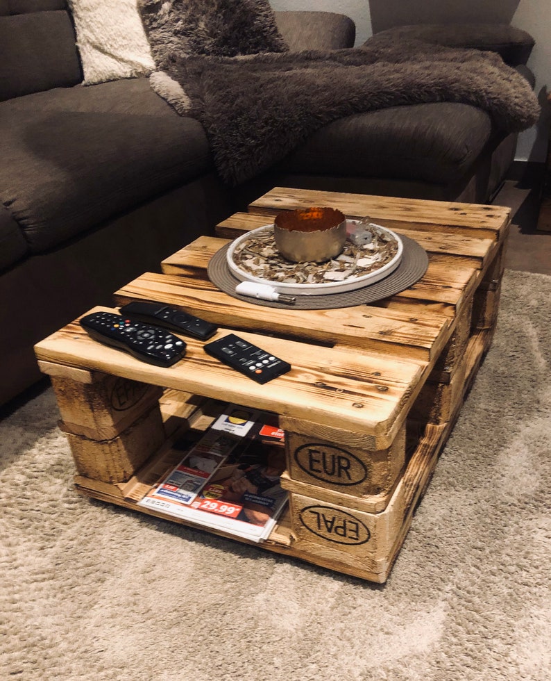 Puede incluir: Una mesa de centro de madera hecha de palets reutilizados. La mesa tiene un aspecto r&uacute;stico y desgastado y presenta las marcas "EUR" y "EPAL". Dos mandos a distancia est&aacute;n sobre la superficie superior de la mesa, y una revista est&aacute; metida en el estante inferior.