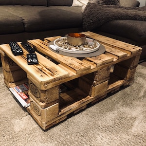 Puede incluir: Una mesa de centro de madera r&uacute;stica hecha de palets reutilizados. La mesa tiene un acabado de madera natural y presenta un plato decorativo con un portavelas en la parte superior. La mesa est&aacute; sentada sobre una alfombra de pelo marr&oacute;n claro.
