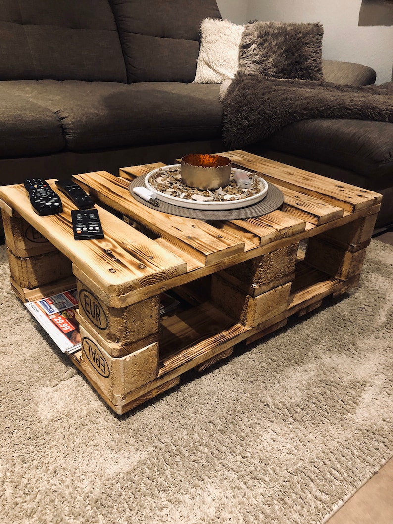 Puede incluir: Una mesa de centro de madera r&uacute;stica hecha de palets reutilizados. La mesa tiene un acabado de madera natural y presenta un plato decorativo con un portavelas en la parte superior. La mesa est&aacute; sentada sobre una alfombra de pelo marr&oacute;n claro.
