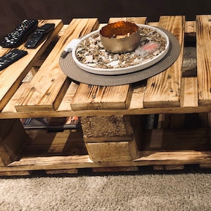 Puede incluir: Una mesa de centro de madera r&uacute;stica hecha de palets reutilizados. La mesa tiene un acabado de madera natural y presenta una bandeja decorativa con un cuenco de metal y relleno blanco. La mesa tiene un estante debajo para almacenamiento.