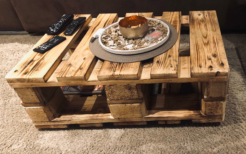 Puede incluir: Una mesa de centro de madera r&uacute;stica hecha de palets reutilizados. La mesa tiene un acabado de madera natural y presenta una bandeja decorativa con un cuenco de metal y relleno blanco. La mesa tiene un estante debajo para almacenamiento.