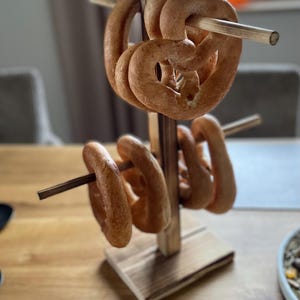 Könnte beinhalten: Ein hölzerner Brezelhalter mit mehreren gebackenen Brezeln. Der Halter besteht aus hellbraunem Holz und hat horizontale Dübel zur Aufnahme der Brezeln. Die Brezeln sind goldbraun und haben eine klassische Brezelform.