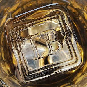 May include: A top-down view of a rocks glass filled with amber liquid and a large ice cube. The ice cube has the letters "TB" embossed on it. The glass sits on a dark, textured surface.
