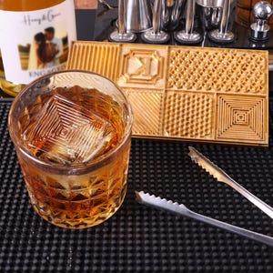 Może przedstawiać: Złota metalowa tacka na kostki lodu z różnymi wzorami. Taca znajduje się na czarnej powierzchni z szklanką whisky i kostkami lodu. Kostki lodu są kwadratowe i mają geometryczny wzór.