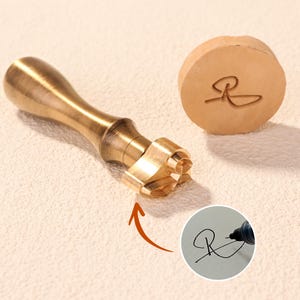 Puede incluir: Un sello de metal de latón con un mango de madera y un cabezal de sello detallado. El sello está junto a un trozo redondo de cuero con la letra "R" grabada. Una pequeña imagen circular muestra la letra "R" siendo escrita.