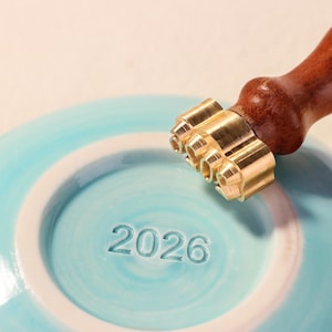Puede incluir: Un sello de lat&oacute;n con un mango de madera se coloca sobre una superficie de cer&aacute;mica azul claro. El sello est&aacute; a punto de imprimir el n&uacute;mero "2026" en la cer&aacute;mica. La cer&aacute;mica tiene un borde blanco y un dise&ntilde;o en espiral.