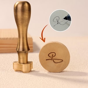 May include: A brass wax seal stamp with a wooden handle. The stamp has a circular base with a custom signature design. An inset shows the signature being written with a black marker. The stamp is used for sealing documents or adding a personalized touch.