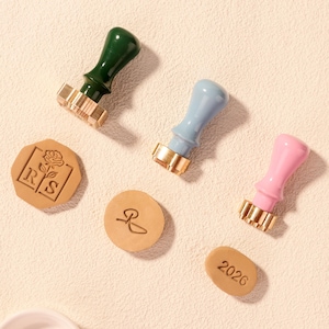 May include: Three wax seal stamps with wooden handles in green, blue, and pink, alongside three embossed wax seals. The wax seals feature a rose, a monogram, and the year 2026. The stamps have gold-colored metal stamp heads.