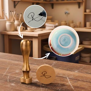 Könnte beinhalten: Ein messingfarbener Töpferstempel mit Holzsockel und Signatur. Der Stempel befindet sich neben einer Keramikschale mit blau-türkisfarbener Glasur und derselben Signatur. Das Bild zeigt auch die Signatur, die mit einem schwarzen Stift geschrieben wird.