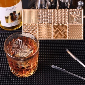 Könnte beinhalten: Ein goldfarbenes Tablett mit neun verschiedenen strukturierten Quadraten. Das Tablett befindet sich auf einer schwarzen Oberfläche mit einem Glas Whisky und Eiswürfeln. Die Eiswürfel haben eine quadratische Form mit einem strukturierten Design.