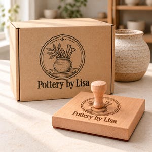 Könnte beinhalten: Ein brauner Karton und ein quadratischer Holzstempel, beide mit dem Pottery by Lisa Logo versehen, das eine Topfpflanze und Töpferwerkzeuge in einem kreisförmigen Rahmen zeigt. Rechts neben dem Karton steht eine gesprenkelte Keramikvase.