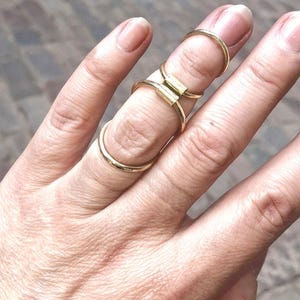 May include: A gold-toned metal finger ring featuring a multi-band design that spans across two knuckles. The piece consists of a thin circular band at the base, a central rectangular bridge connector, and a thinner circular band at the top knuckle.