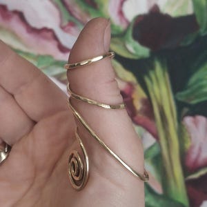 Puede incluir: Una férula para dedo de metal dorado con un diseño de espiral continua que envuelve el pulgar. La estructura consiste en un alambre fino y pulido que se enrolla varias veces, terminando en un detalle de espiral en la base del pulgar.
