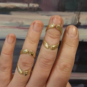 Könnte beinhalten: Zwei goldene Ringe mit einer Lücke in der Mitte, die an zwei Fingern einer Hand getragen werden. Die Ringe sind so konzipiert, dass sie am mittleren Knöchel des Fingers getragen werden.