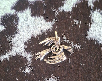 May include: A silver-toned pendant featuring a stylized design of two hands and a spiral. The pendant has a small loop at the top for a chain. The background is a brown and white spotted pattern.