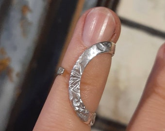 May include: A silver-toned finger ring with a textured, crescent-shaped design. The ring is worn on a finger, and the design features an etched pattern. The ring appears to be adjustable.