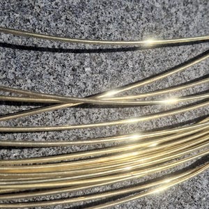 Può includere: Primo piano di diversi fili sottili e lucenti color oro disposti in un fascio. I fili sono appoggiati su una superficie grigia strutturata. I fili riflettono la luce, creando riflessi luminosi.