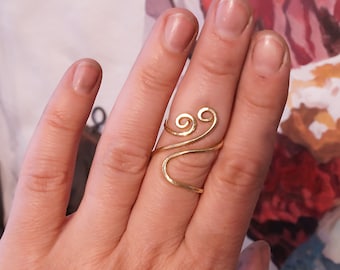May include: A gold-toned, wire ring with two swirls on the top. The ring is worn on a finger.