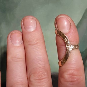 Puede incluir: Un anillo dorado con un diseño abierto, usado en el dedo índice de una mano.