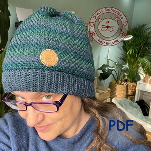 Könnte beinhalten: Eine handgestrickte Mütze in Türkis-, Blau- und Grüntönen. Die Mütze hat einen umgeschlagenen Saum und einen runden Lederaufnäher. Das Bild enthält auch den Text "PDF".