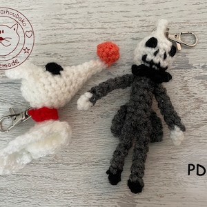 Könnte beinhalten: Zwei gehäkelte Schlüsselanhänger, einer weiß mit rotem Halsband und schwarzer Nase, der andere grau mit schwarzem Totenkopf und Fliege. Beide Schlüsselanhänger haben silberne Schlüsselringe. Der Text "PDF" ist in der rechten unteren Ecke des Bildes sichtbar.