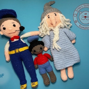 Puede incluir: Tres muñecas de crochet, una muñeca vestida de azul y amarillo con un sombrero azul, una muñeca vestida de azul y rojo con un sombrero azul, y una muñeca vestida de azul con barba blanca y un sombrero gris. Las muñecas están sobre un fondo azul. La imagen también incluye un círculo rojo con un gato blanco y el texto "Dre's Saihoubako homemade".