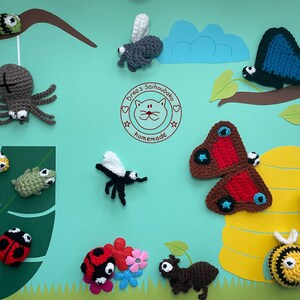 Könnte beinhalten: Eine Sammlung von gehäkelten Insektenspielzeugen, darunter eine Spinne, eine Raupe, ein Frosch, ein Marienkäfer, ein Schmetterling, eine Biene und eine Ameise. Die Spielzeuge werden auf einem blauen Hintergrund mit grünen Blättern und einem gelben Bienenstock ausgestellt.