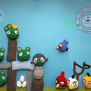 Könnte beinhalten: Gehäkelte Angry Birds Charaktere und Strukturen vor blauem Hintergrund. Die Szene zeigt grüne Schweineköpfe, einen blauen Vogel und verschiedene rote, weiße, gelbe und schwarze Vögel. Auch eine Schleuder ist vorhanden.