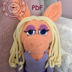 Könnte beinhalten: Handgemachte Häkelpuppe eines Schweinecharakters mit blonden Garnhaaren, blauen Augen und einem lila Kleid. Die Puppe hat große Ohren und ist pfirsichfarben. Der Text "PDF" ist in der oberen rechten Ecke sichtbar.