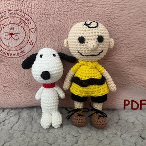 Könnte beinhalten: Gehäkelte Snoopy- und Charlie Brown-Puppen. Snoopy ist weiß mit schwarzen Ohren und einem roten Kragen. Charlie Brown trägt ein gelbes Hemd mit einem schwarzen Zickzack und braunen Schuhen. Das Wort "PDF" ist rot.
