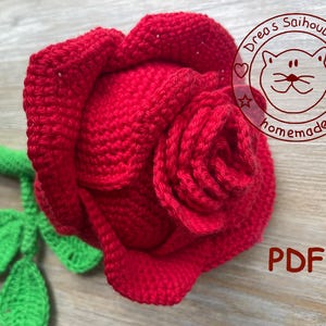 Könnte beinhalten: Eine handgehäkelte rote Rose mit grünem Stiel und Blättern. Die Rose steht in voller Blüte, mit detaillierter Blütenblattarbeit. Das Bild enthält den Text "PDF" und ein kreisförmiges Logo mit dem Text "Drea's Saihoubako homemade".