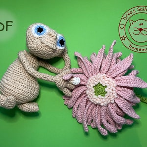 Könnte beinhalten: Ein gehäkeltes Amigurumi-Spielzeug eines grauen Außerirdischen, der eine rosa gehäkelte Blume hält. Der Außerirdische hat große blaue Augen und einen kleinen Mund. Die Blume hat eine grüne Mitte und rosa Blütenblätter. Der Hintergrund ist eine grüne Oberfläche. Das Bild enthält auch einen braunen Kreis mit dem Text "Drea's Saihoubako homemade" in weiß.