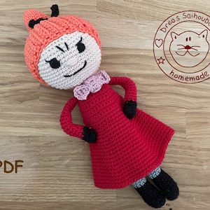 May include: A crocheted doll with a red dress, orange hair, and a black and white face. The doll is lying on a wooden surface. The text "Drea's Saihoubako homemade" is visible in a circle with a cat face in the upper right corner of the image.